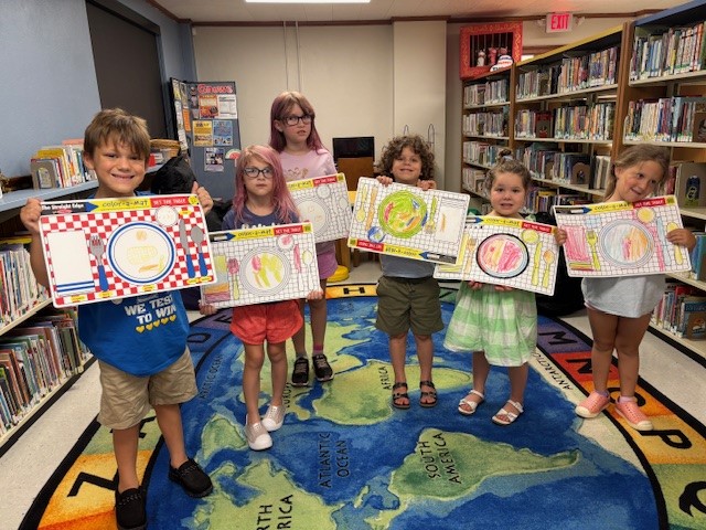 Six children holding colorful certificates while standing on a world map carpet in a library.